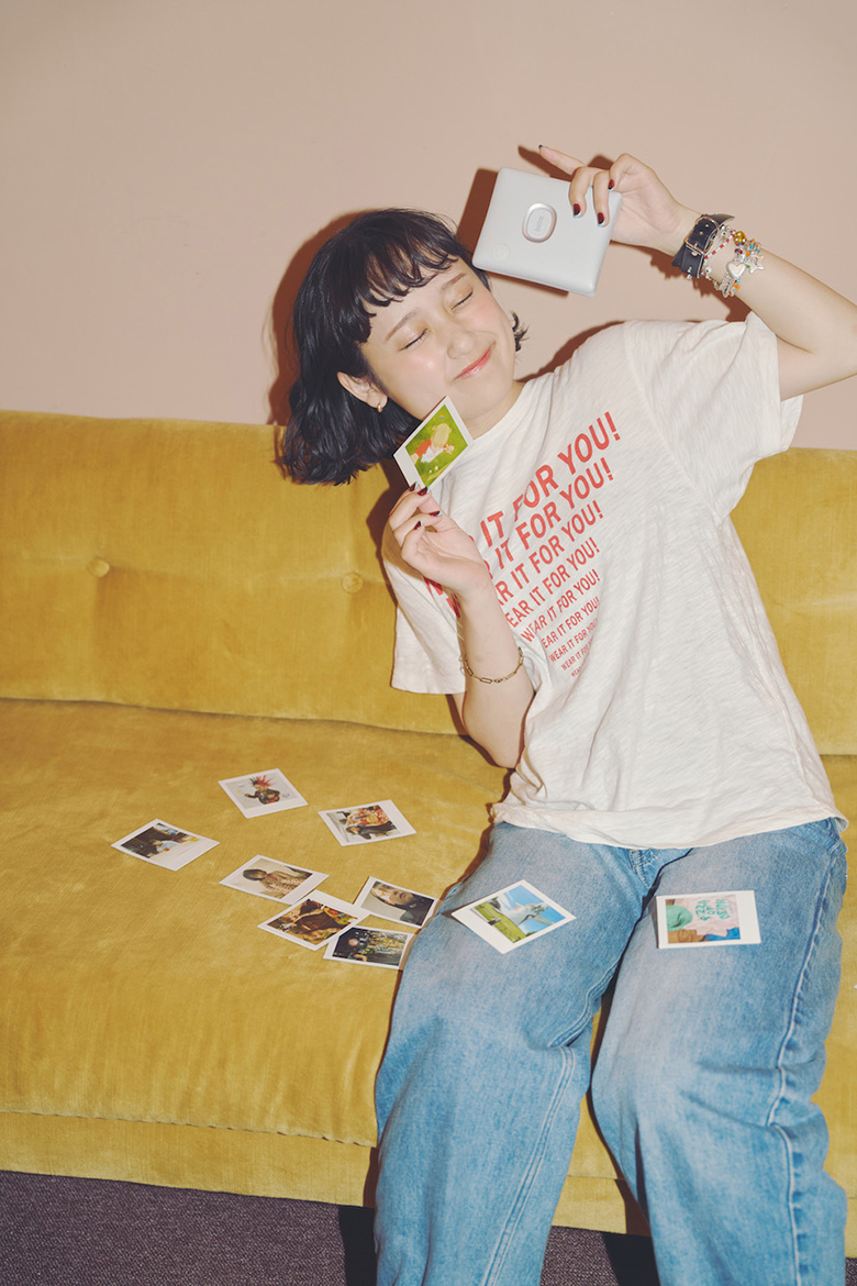 ソファに座ったなちさんがSQUARE Linkとチェキプリントを顔の横で持って目を閉じている写真